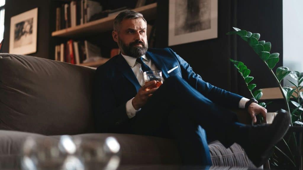 Man with a gray beard in a suit sitting on a couch, holding a glass of amber liquid.