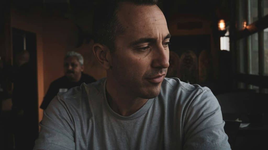Man in a gray shirt sits indoors, looking down thoughtfully with a blurry person in the background.