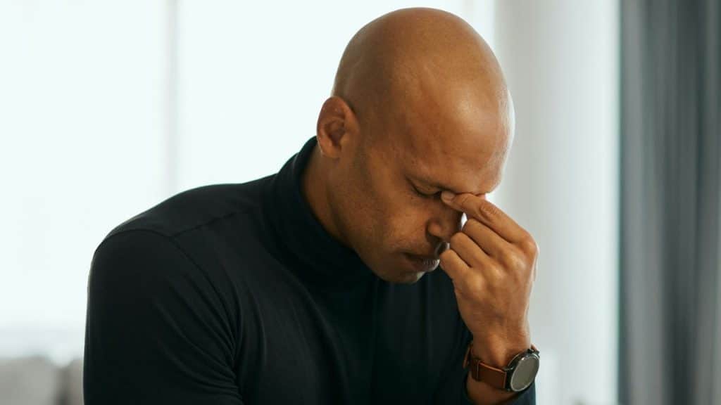 Bald man in a black turtleneck sitting with his eyes closed and hand on his forehead.