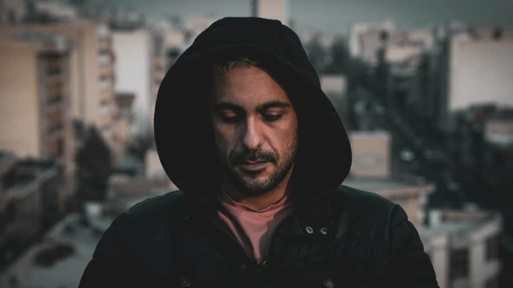 Man in a black hooded jacket looking down sadly, with a blurry city background.