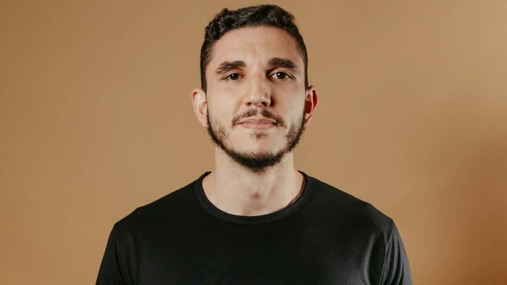 Man with dark hair and a short beard in a black t-shirt against a tan background.