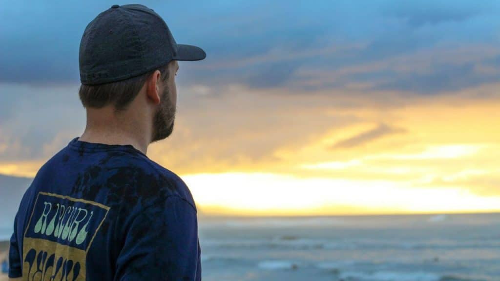 Man in a dark shirt and baseball cap looking out at the ocean at sunset.
