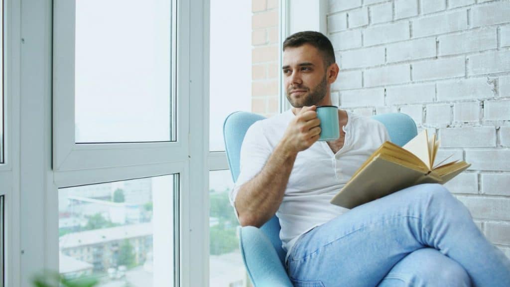 Man in a white shirt and jeans sitting in an armchair, reading a book and holding a mug.