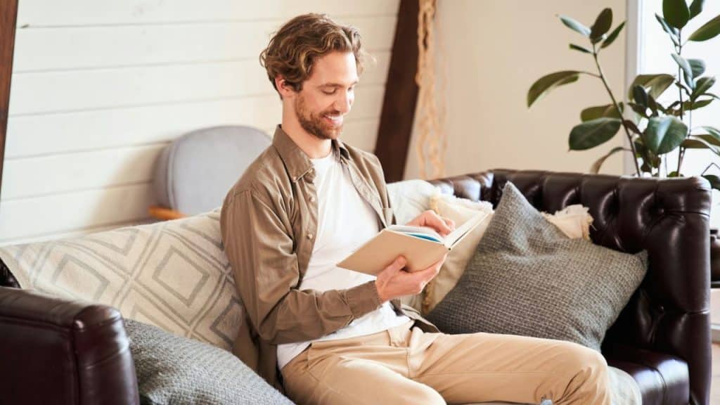 Smiling man with wavy hair sitting on a couch, reading a light-colored book.
