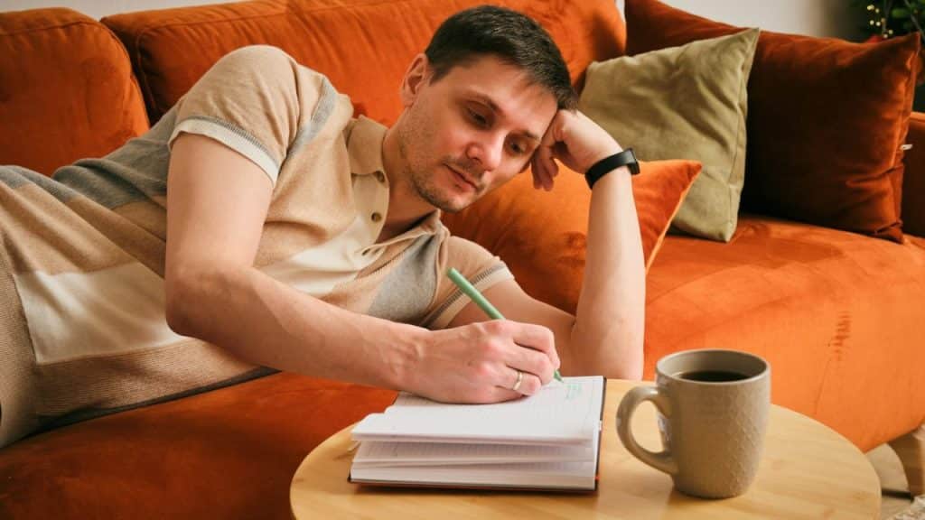 Man reclining on an orange couch, writing in a notebook next to a mug of coffee.