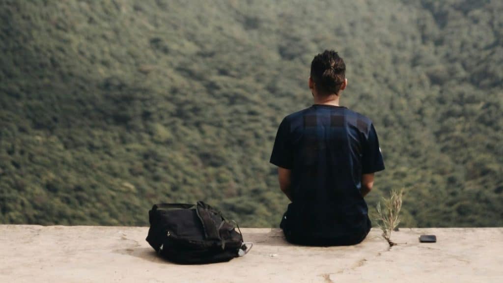 Man seated with his back to the camera, looking out at a densely forested hillside, with a backpack beside him.