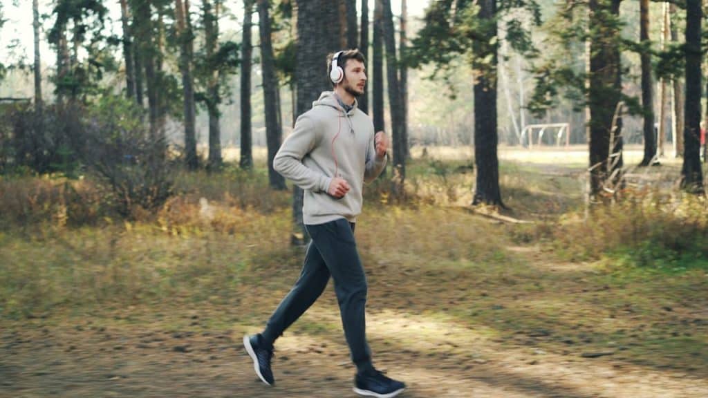 Man wearing a light hoodie and headphones running through a wooded area.