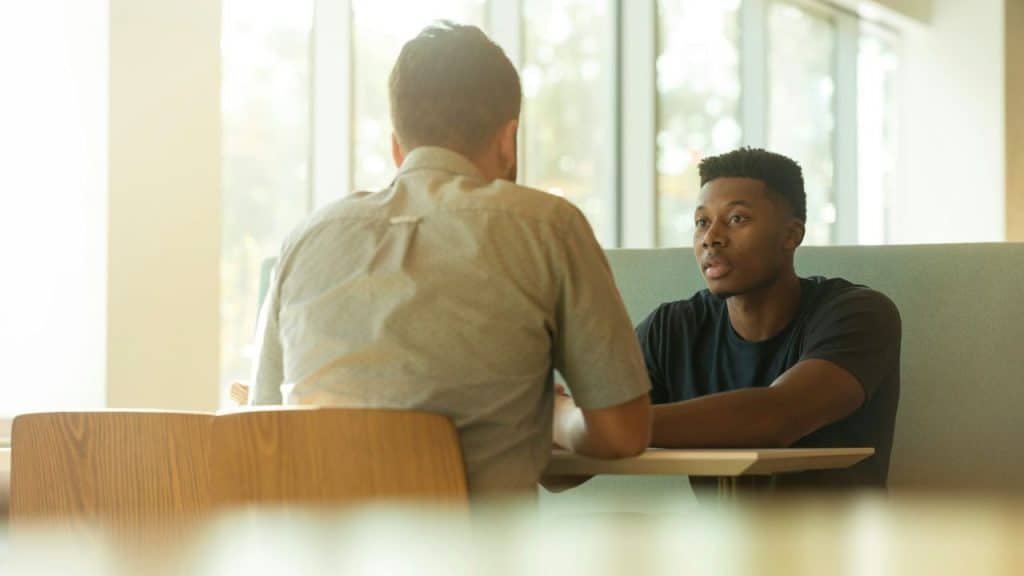 Two men seated at a table, one with his back to the camera, the other listening intently.