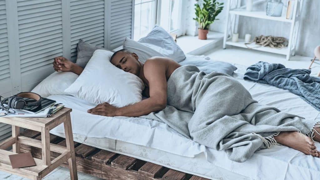 Man sleeping on his side under a light blanket in a bright bedroom.