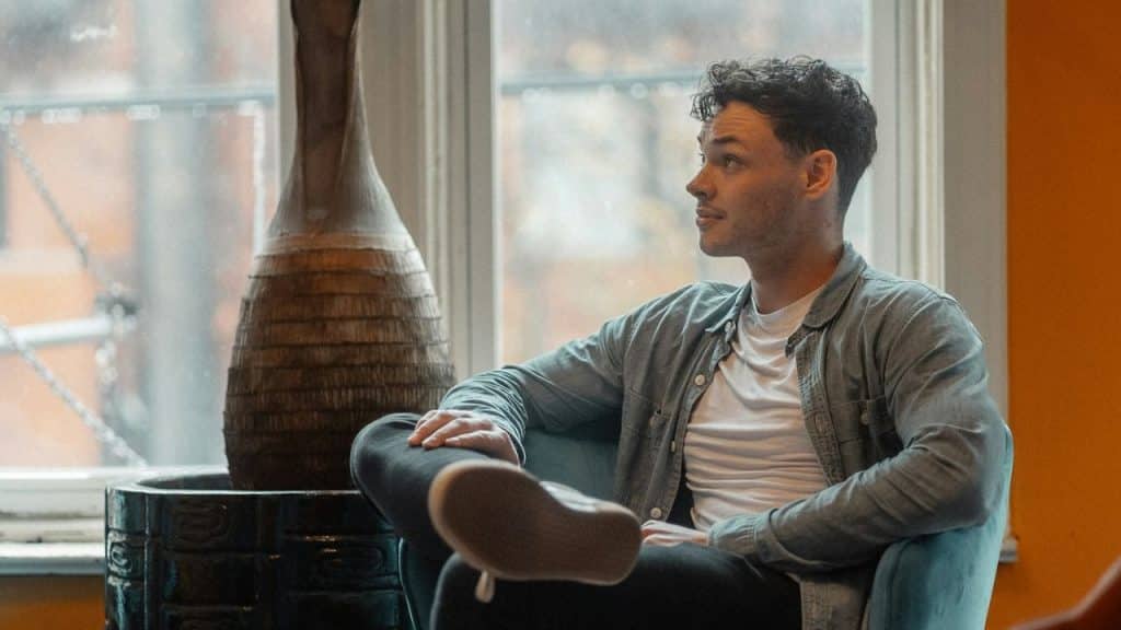 Man sitting in a chair with his legs crossed, looking out a window with a large vase next to him.
