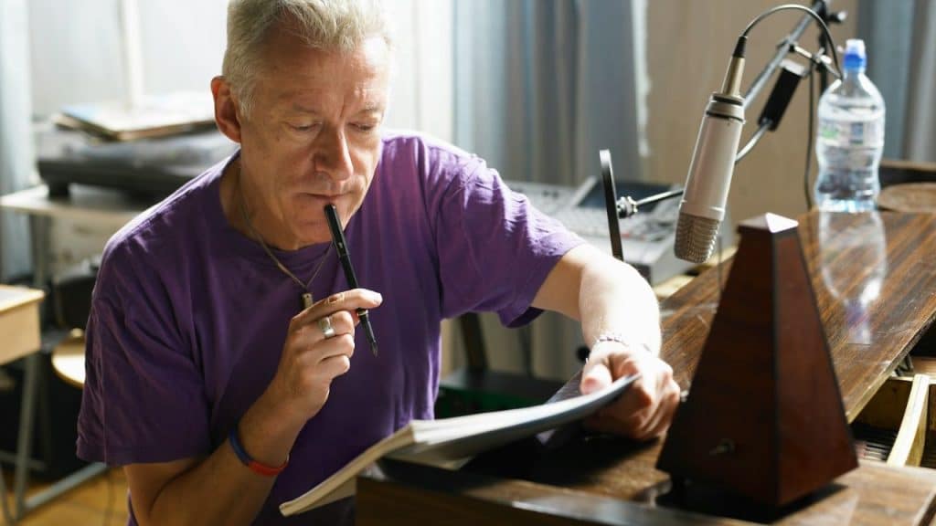 Older man in a purple shirt looking intently at a notebook, holding a pen to his mouth, with a microphone and metronome nearby.