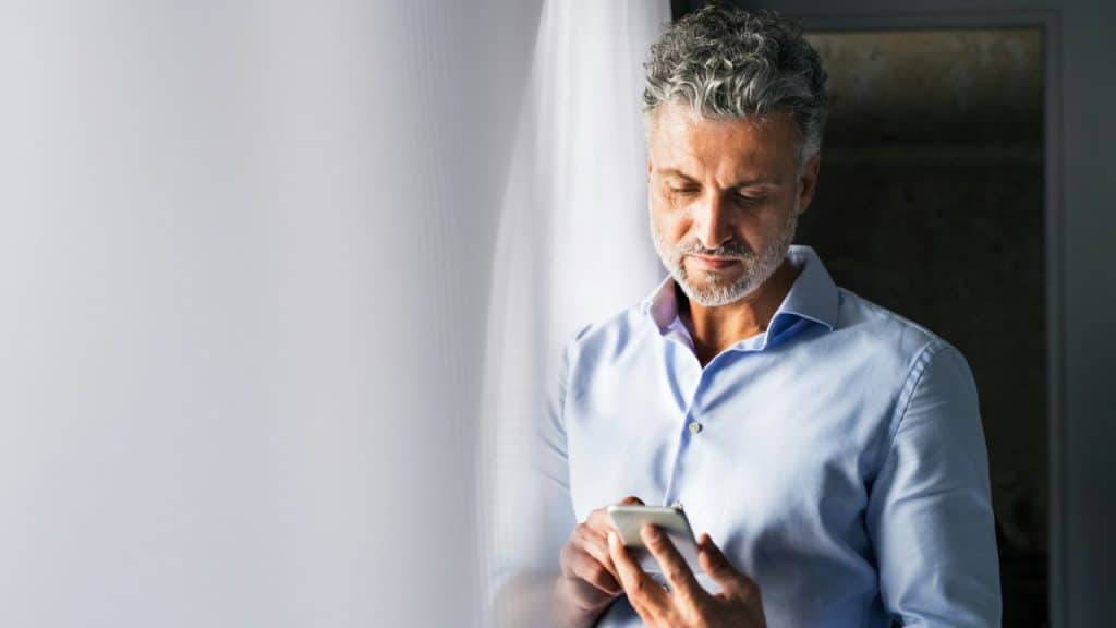 Middle-aged man with gray hair and a beard, wearing a blue shirt, looking intently at his smartphone.