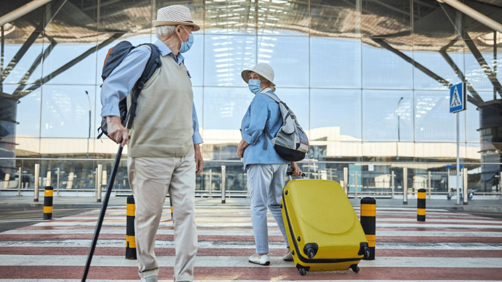 An older couple wearing masks and hats stands outside an airport with travel luggage.