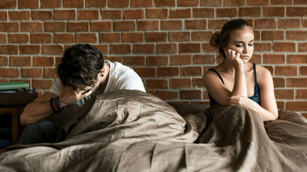 A distressed couple sits apart in bed, facing away from each other with sad expressions.