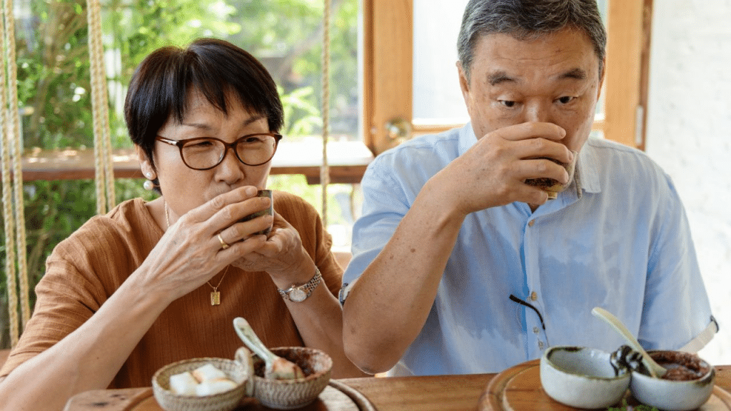 An older Asian couple drinks from small cups together in a cafe.