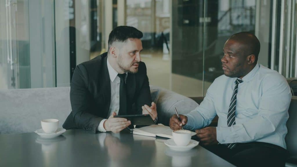 Two businessmen sitting at a table discussing, with coffee cups and a notebook.