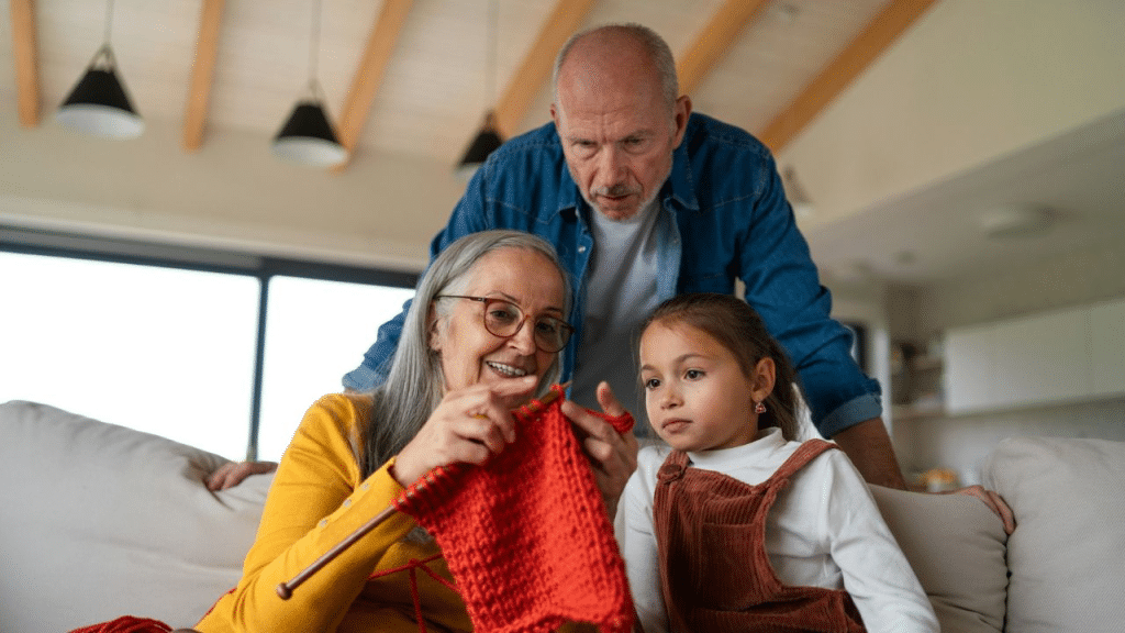 A happy grandmother teaches her young granddaughter to knit as a grandfather watches.