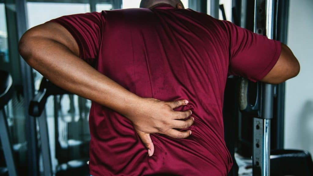 Man in a maroon shirt seen from the back, clutching his lower back in pain at a gym.