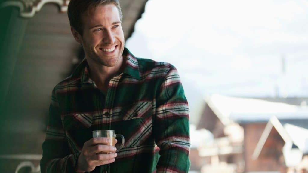 Smiling man in a green and red plaid shirt standing outdoors, holding a glass mug.