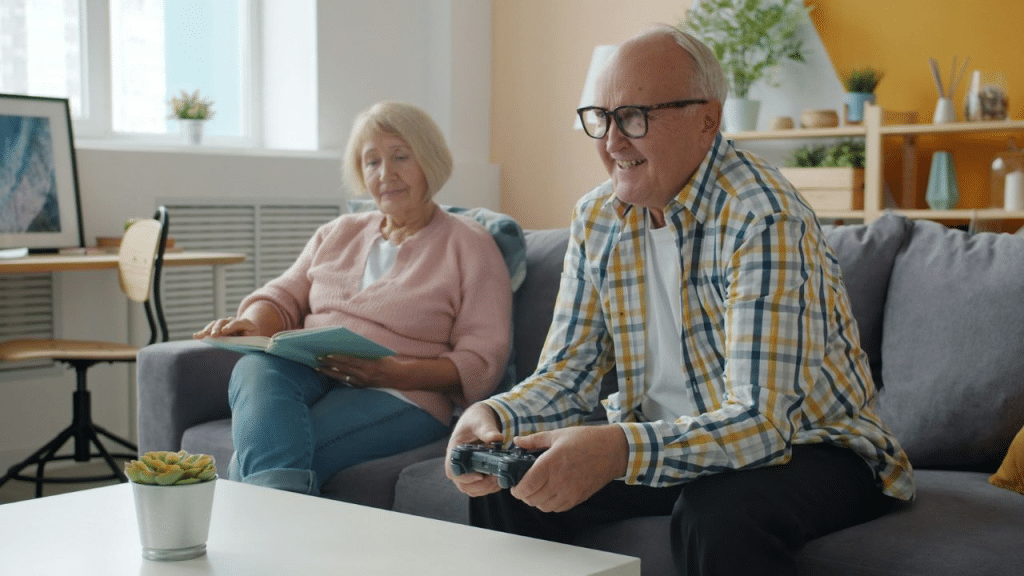 A joyful older man plays a video game while an older woman reads on the couch.
