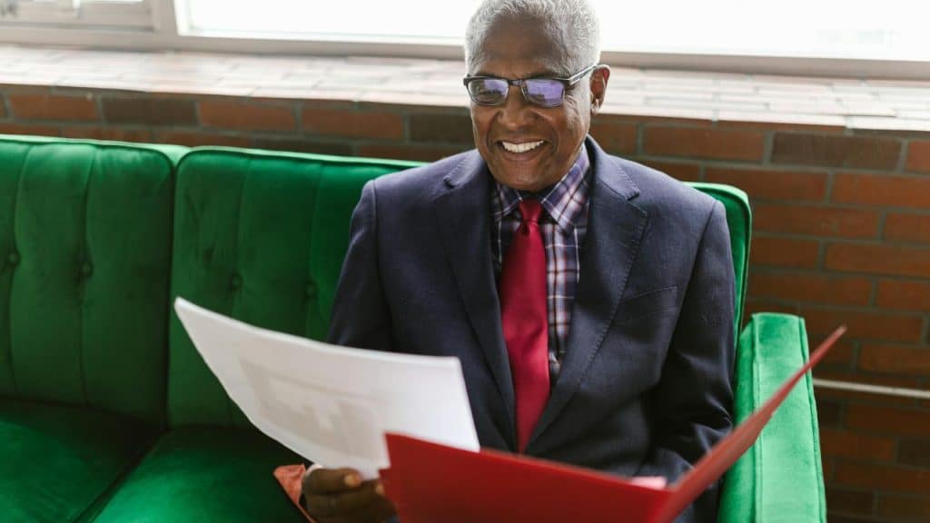 A senior man in a suit smiling while reading documents on a green couch.