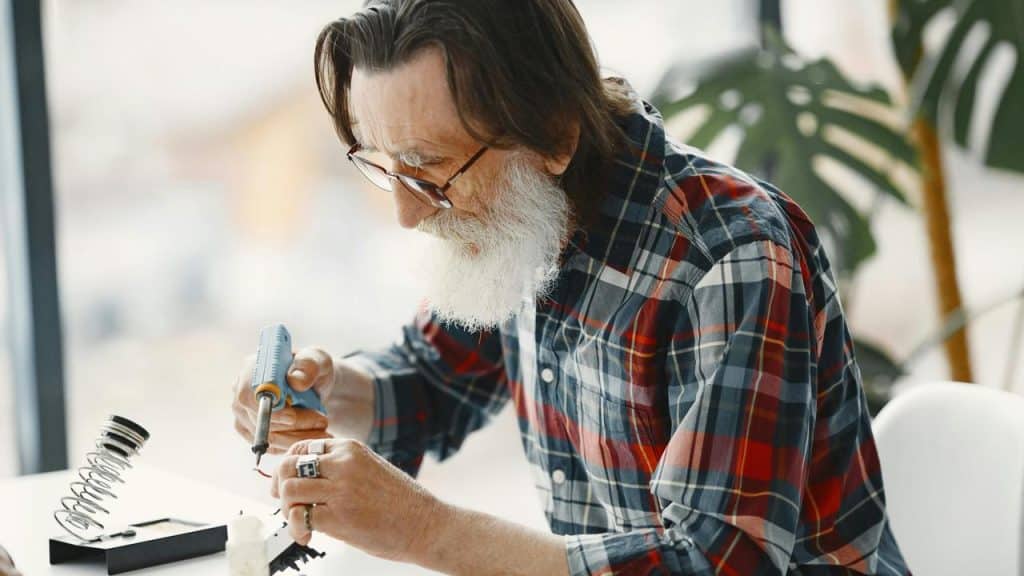 An older man with a beard working with a soldering iron at a table.