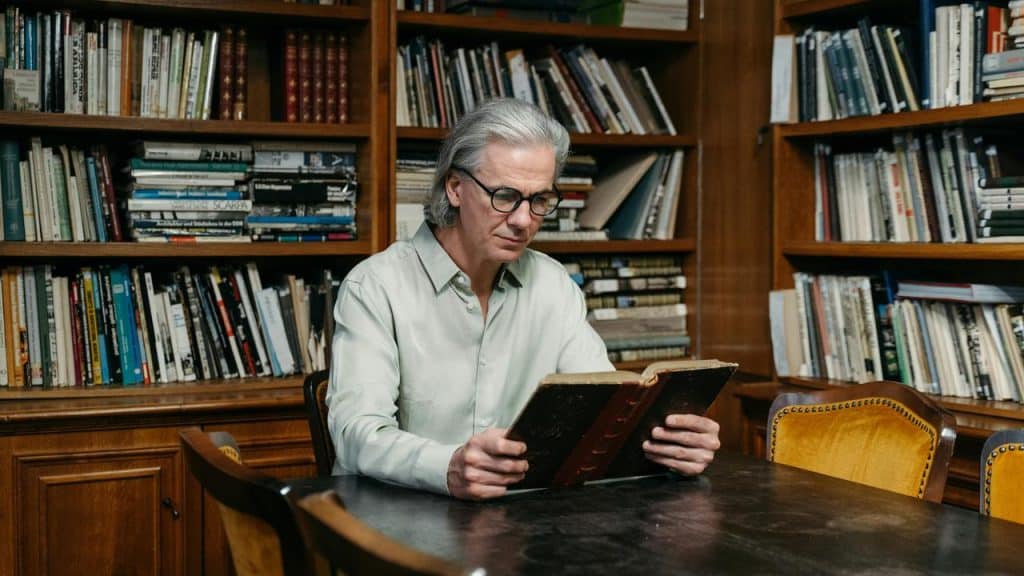 A middle-aged man reading a book at a table in a library.