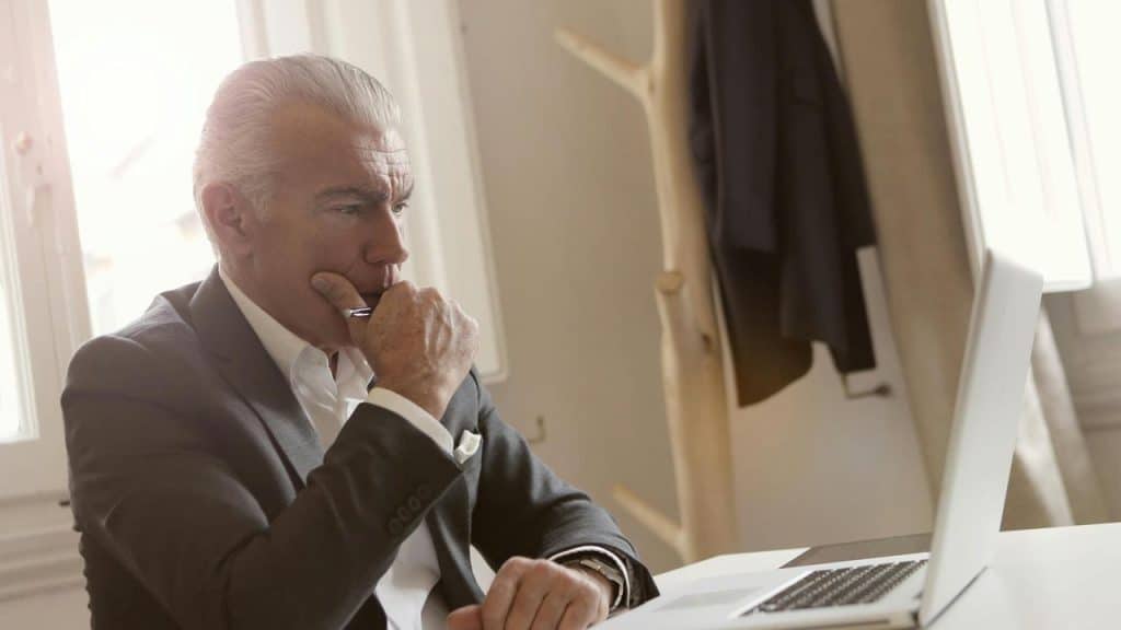 A mature businessman sitting at a desk and thinking while looking at a laptop.