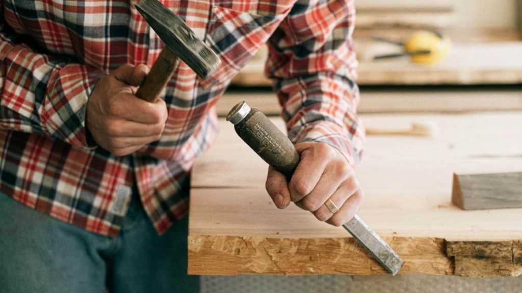 A person wearing a plaid shirt using a hammer and chisel to work on wood.