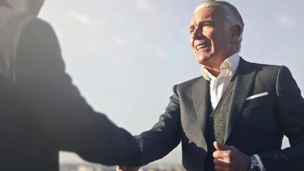 A senior businessman smiling while shaking hands with another person outdoors.