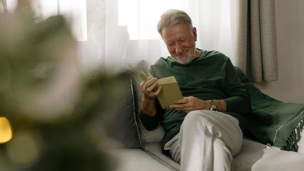 A senior man sitting on a couch smiling while reading a book.