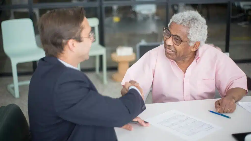 An older man smiling while shaking hands with another man across a table.