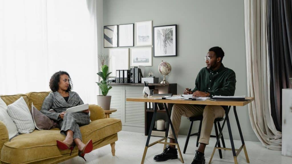 A woman sitting on a couch holding a tablet while talking to a man at a desk.