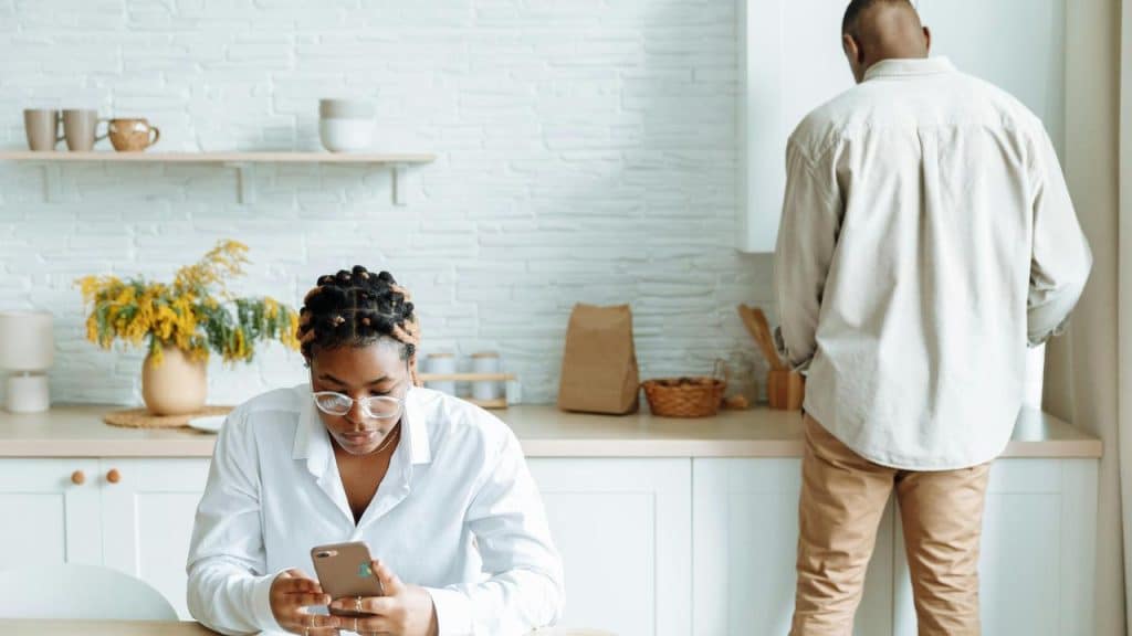 A woman sitting at a table looking at her phone while a man stands facing away in the kitchen.