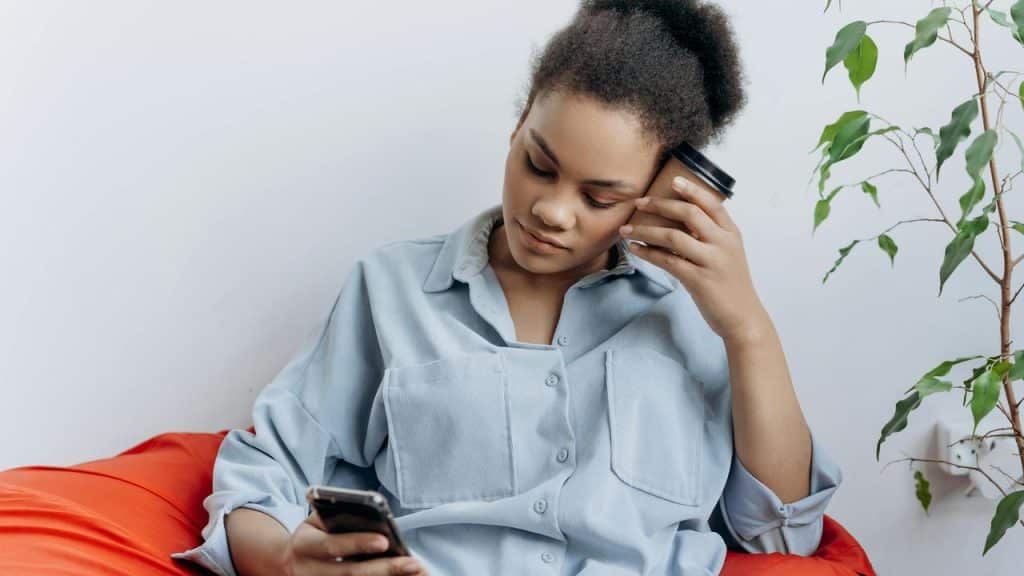 A woman sitting with a cup resting on her head while looking at her phone.