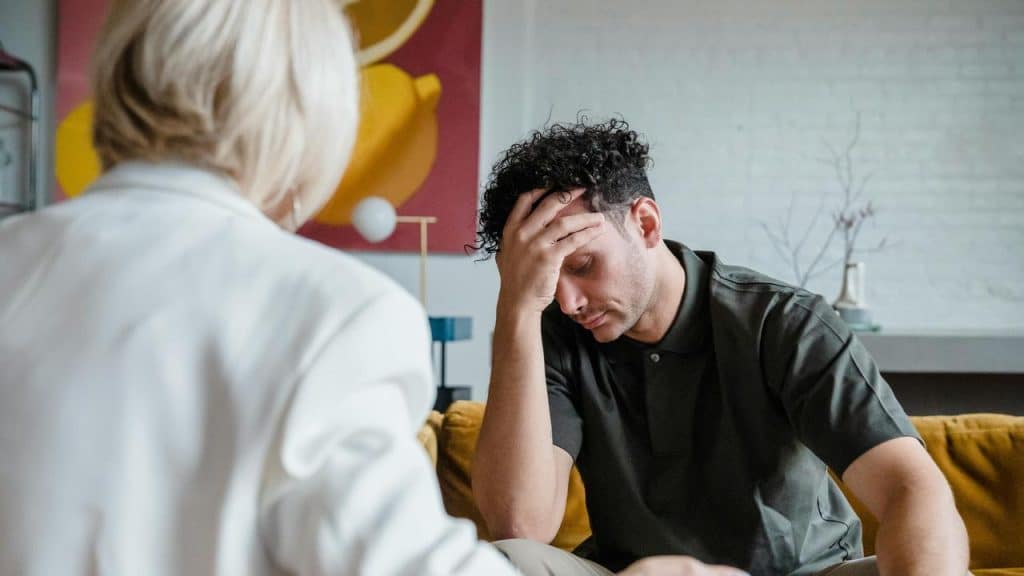 A man sitting on a couch with his head in his hand while another person listens.