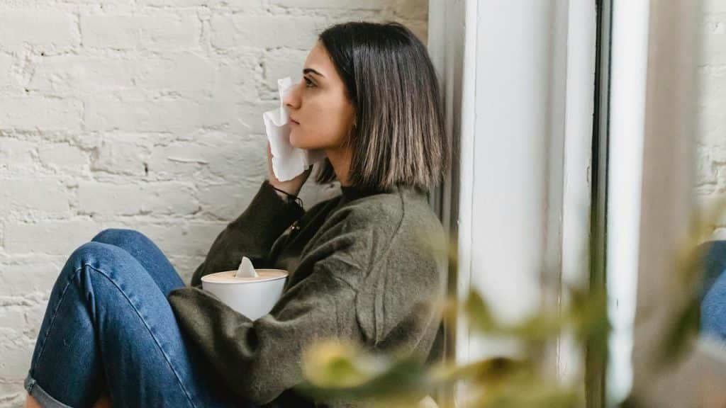A woman sitting by a window holding a tissue to her face with a tissue box on her lap.