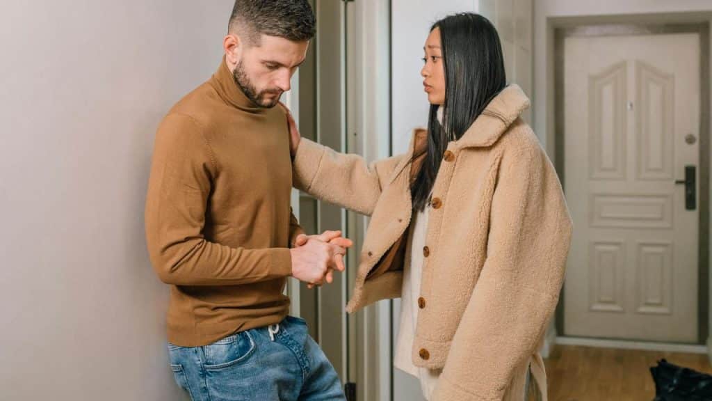 A woman standing with her hand on a man’s shoulder while he looks down with clasped hands.
