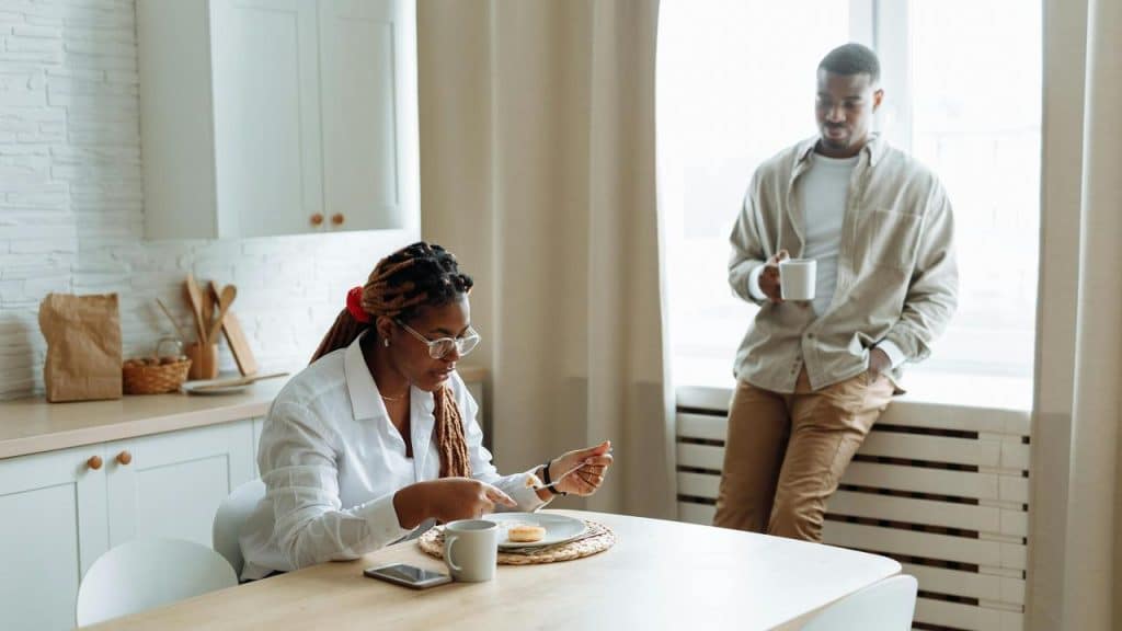 A woman sitting at a kitchen table eating while a man stands nearby holding a mug.