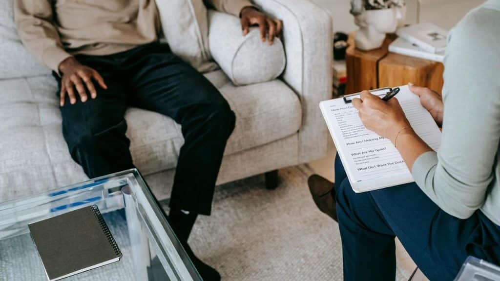 A person sitting on a couch while another person takes notes on a clipboard.