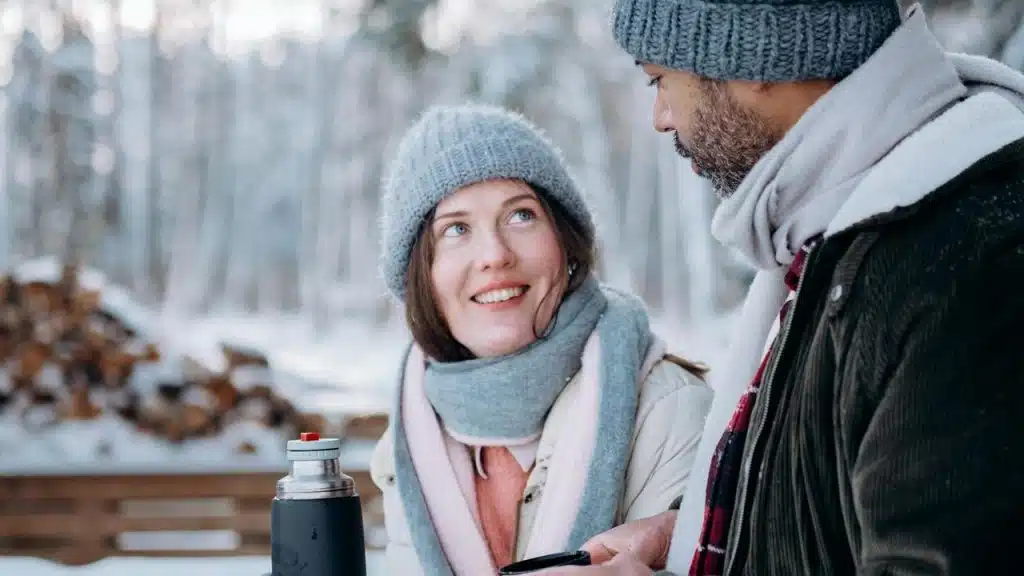 A woman and a man wearing winter clothes talking outdoors in a snowy forest.