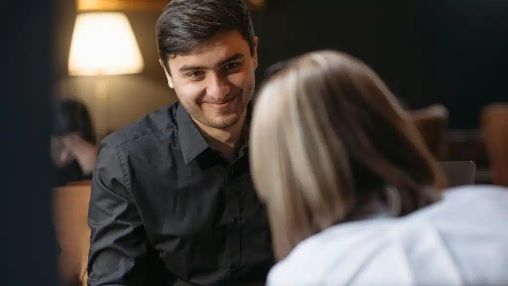 A man smiling while sitting across from a woman in a dimly lit room.