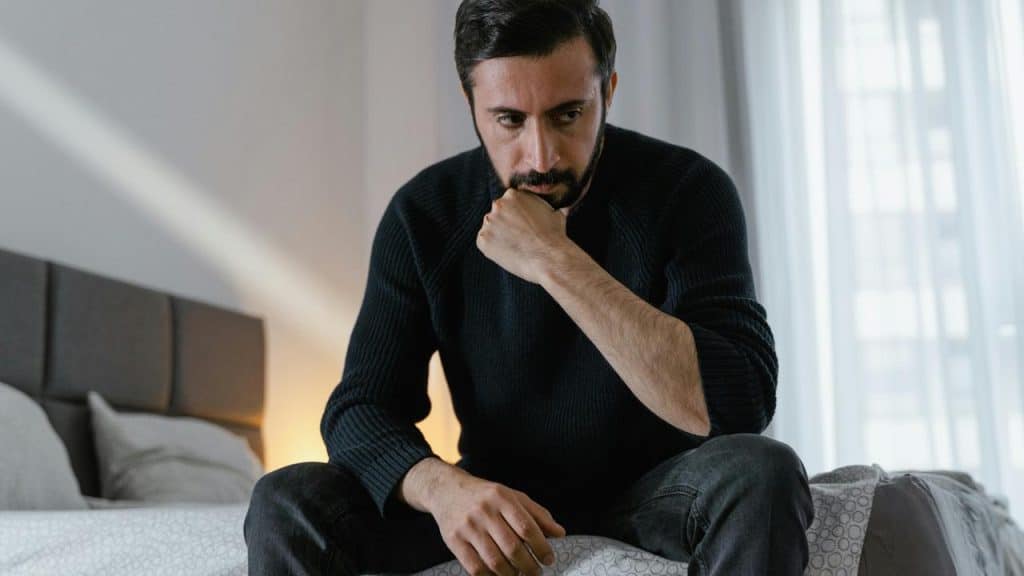 A man sitting on a bed looking thoughtful with his hand on his chin.
