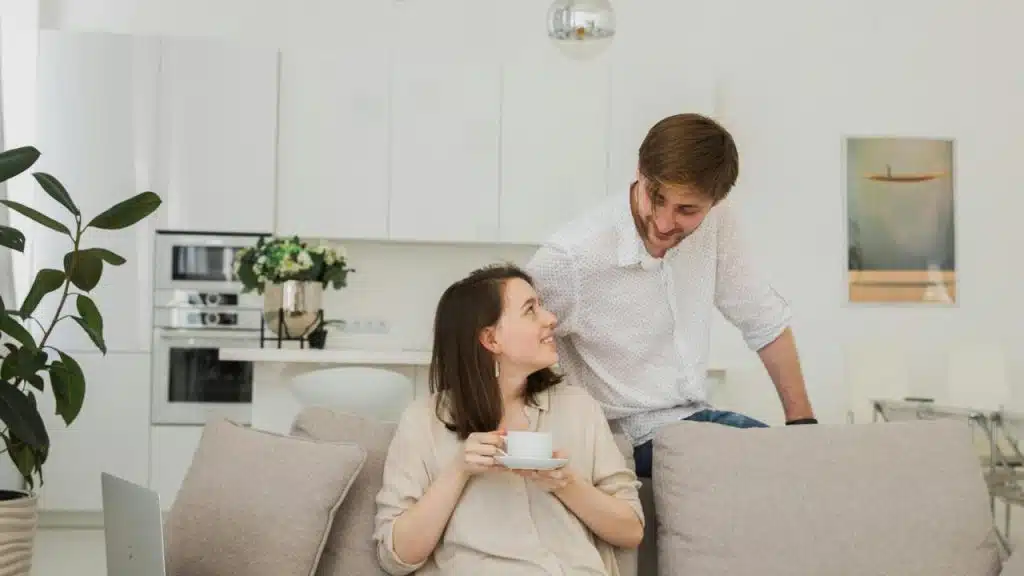 A woman sitting on a couch holding a cup while smiling at a man standing behind her.