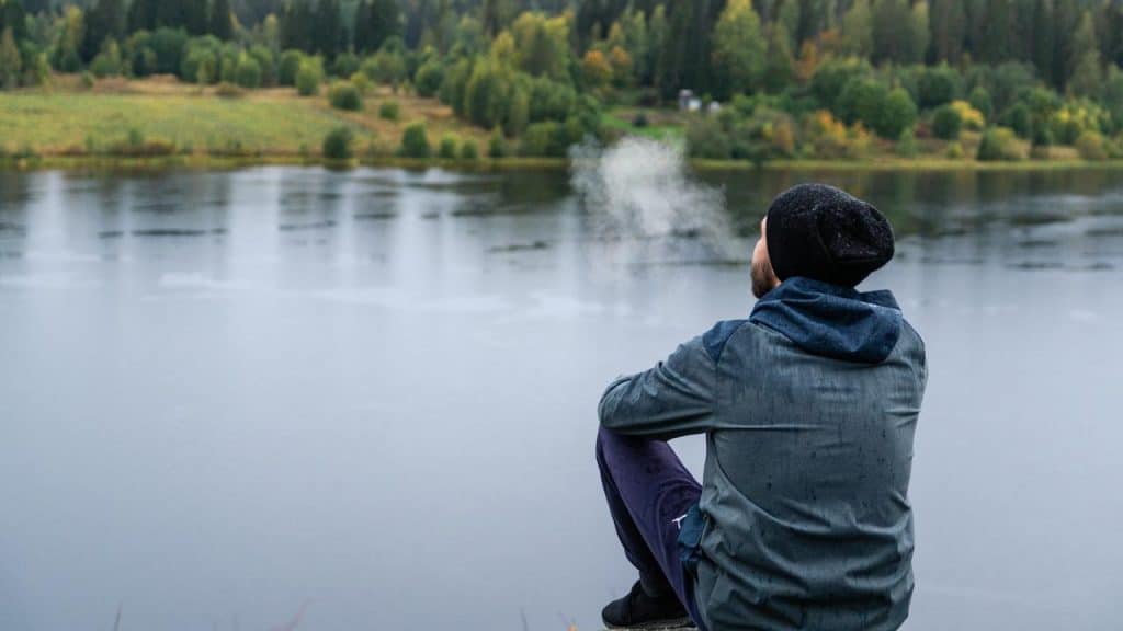 A man wearing a beanie sitting by a lake with trees in the background.