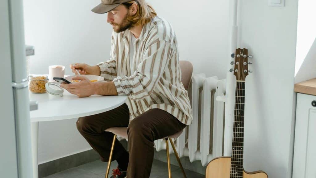 A man eating cereal at a table while looking at his phone with a guitar by the wall.