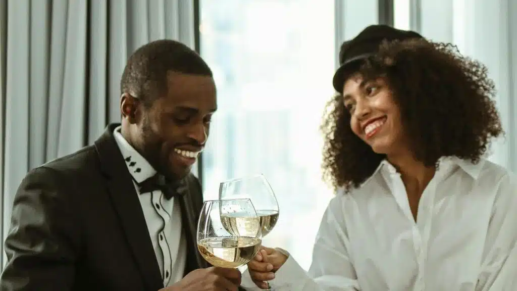 A man in a suit and a woman in a white shirt smiling while clinking wine glasses.