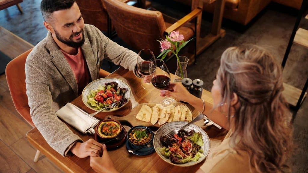 A man and a woman clinking glasses of red wine over a dinner table with salads and bread.