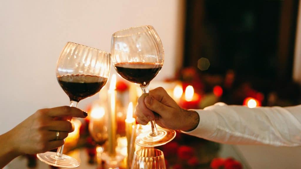 A pair of hands clinking glasses of red wine over a candlelit table.