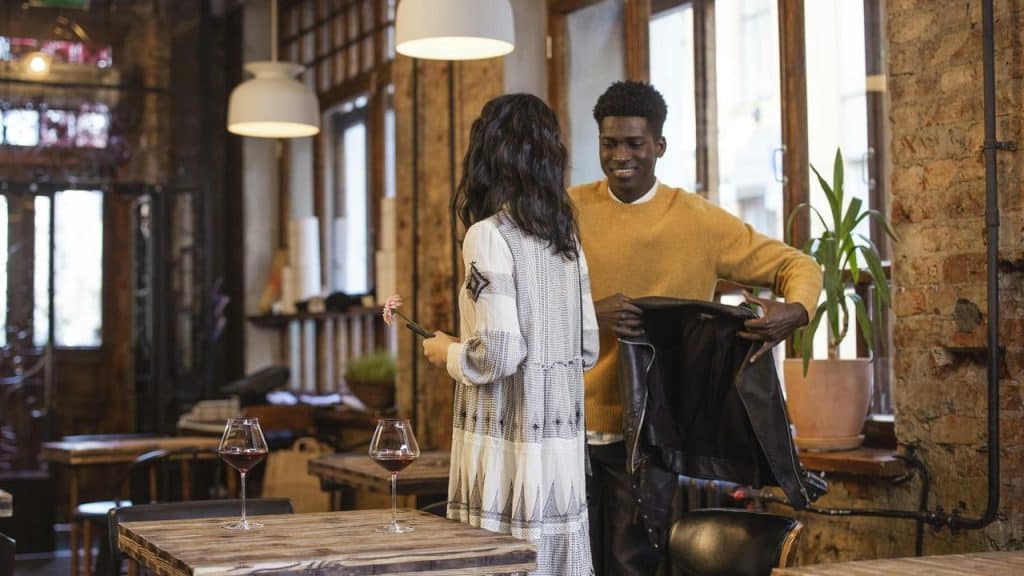 A man in a yellow sweater holding a jacket while smiling at a woman in a café.